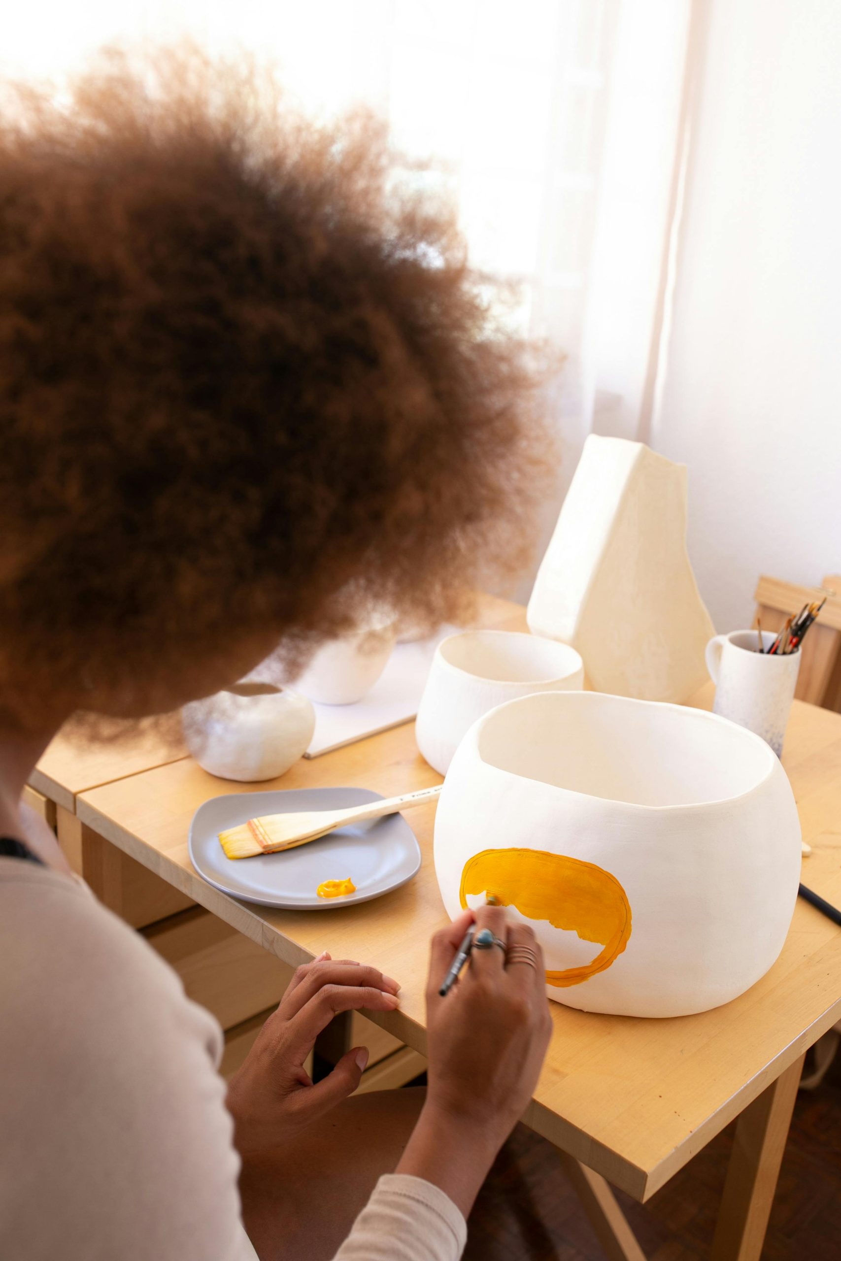 From above back view of crop ethnic female artisan with curly hair sitting at table in workshop and creating pictures with paintbrush on clay tableware